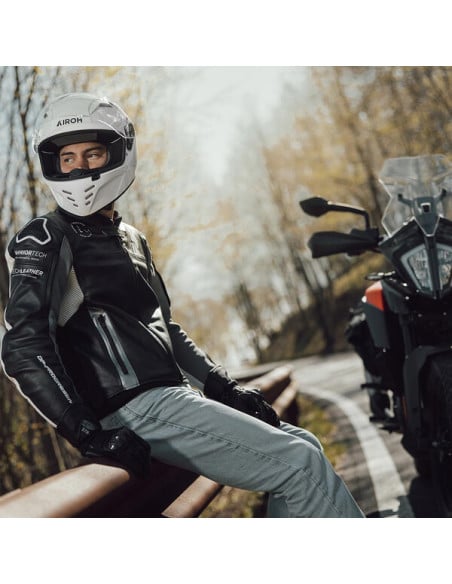 Casque Airoh Conor sur un motard près d’une moto en route forestière, écharpe et gants noirs.